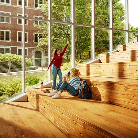 an interior view of two young women , one sitting and one dancing, on wooded steps in front of floor to ceiling windows