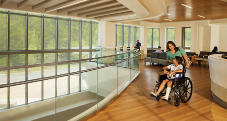 interior photo of a woman pushing a wheelchair with a seated boy, his left leg in a cast, facing a wall of windows bringing sunlight into the room