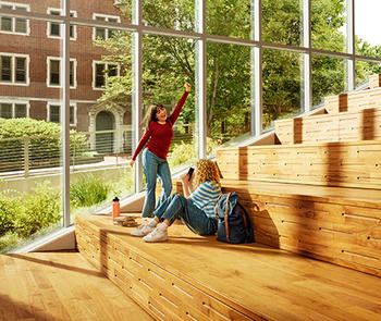 an interior view of two young women , one sitting and one dancing, on wooded steps in front of floor to ceiling windows 