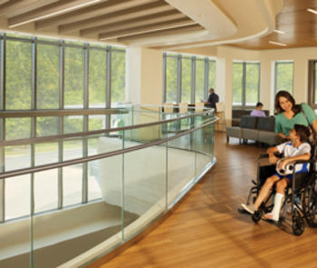 interior photo of a woman pushing a wheelchair with a seated boy, his left leg in a cast, facing a wall of windows bringing sunlight into the room